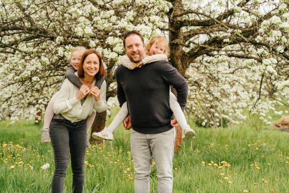 Familie posiert fröhlich im Grünen unter blühendem Baum, Kinder auf den Schultern der Erwachsenen.