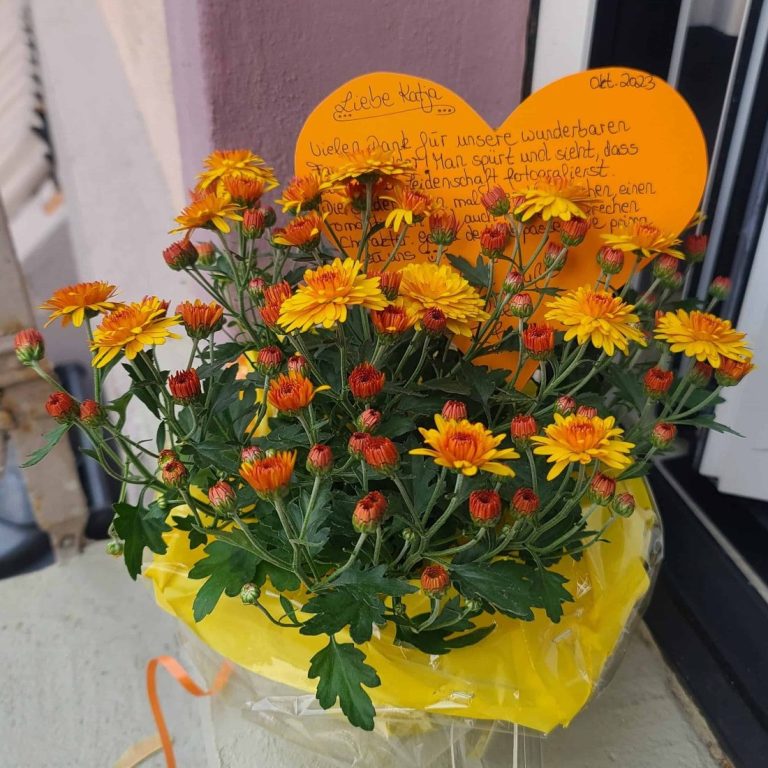 Topf mit leuchtend orange-gelben Chrysanthemen und einer herzförmigen Karte - Geschenk einer Kundin.