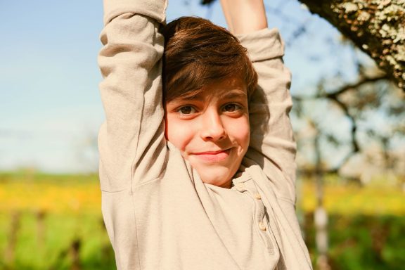 Ein Junge hängt an einem Baumast und lächelt, mit einer Wiese im Hintergrund.