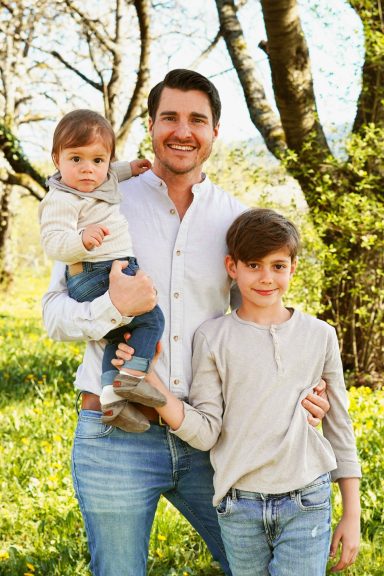 Vater mit zwei Kindern im Freien, umgeben von Bäumen und grünem Gras.