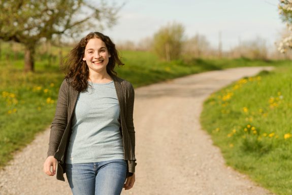 Junge Frau mit braunen Haaren, die auf einem Weg in der Natur lächelt.