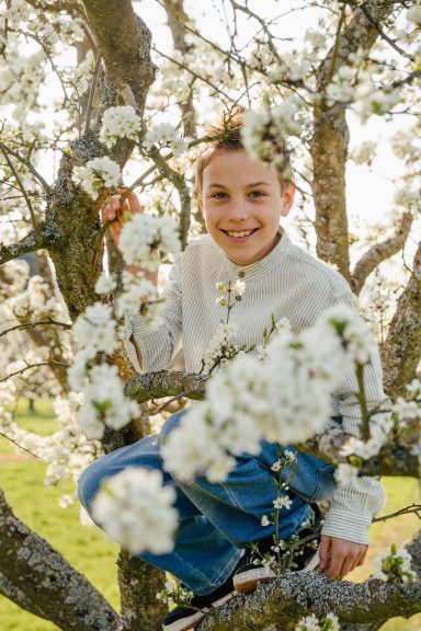 Ein lächelndes Kind sitzt auf einem Baum, umgeben von blühenden weißen Blumen.