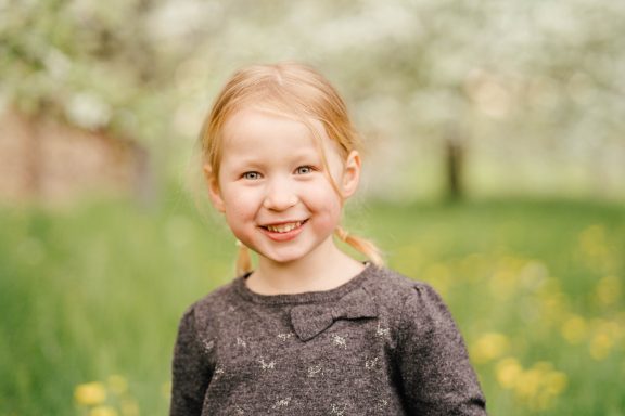 Lächelndes Mädchen mit Zöpfen steht vor einer blühenden Wiese.