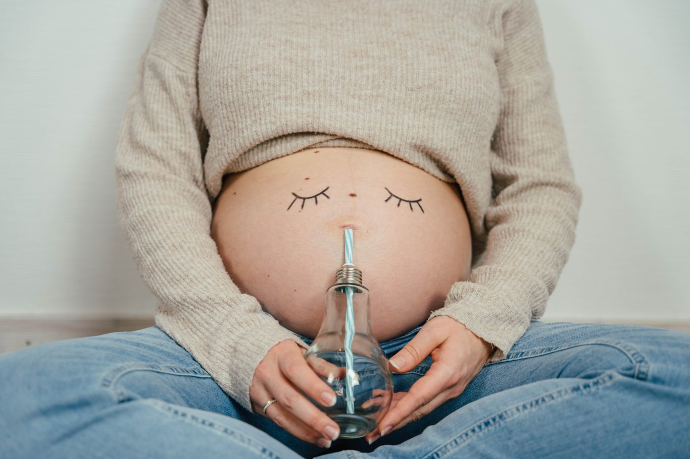 Schwangere Frau, auf deren Bauch Augen gemalt sind und sie ein Wasserglas mit Röhrchen an den Bauchnabel hält.