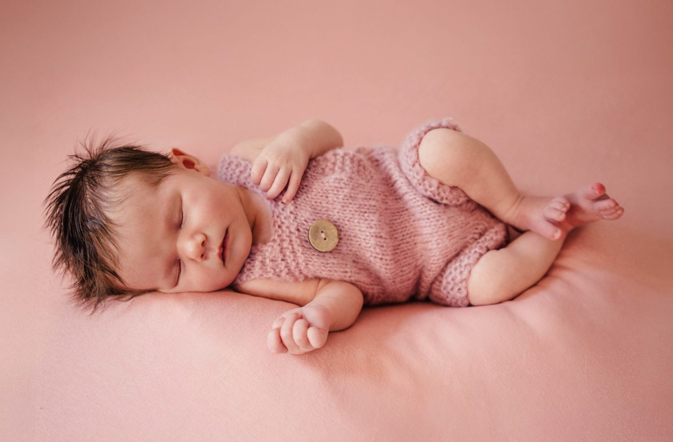 Schlafendes Baby in rosafarbenem Strickoutfit auf einem sanften Untergrund.