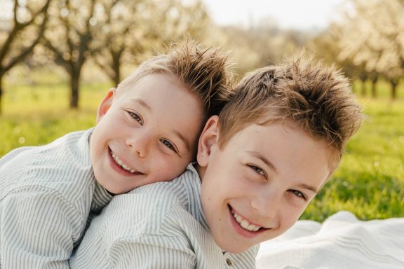 Zwei lächelnde Jungen liegen im Gras, umgeben von blühenden Bäumen.