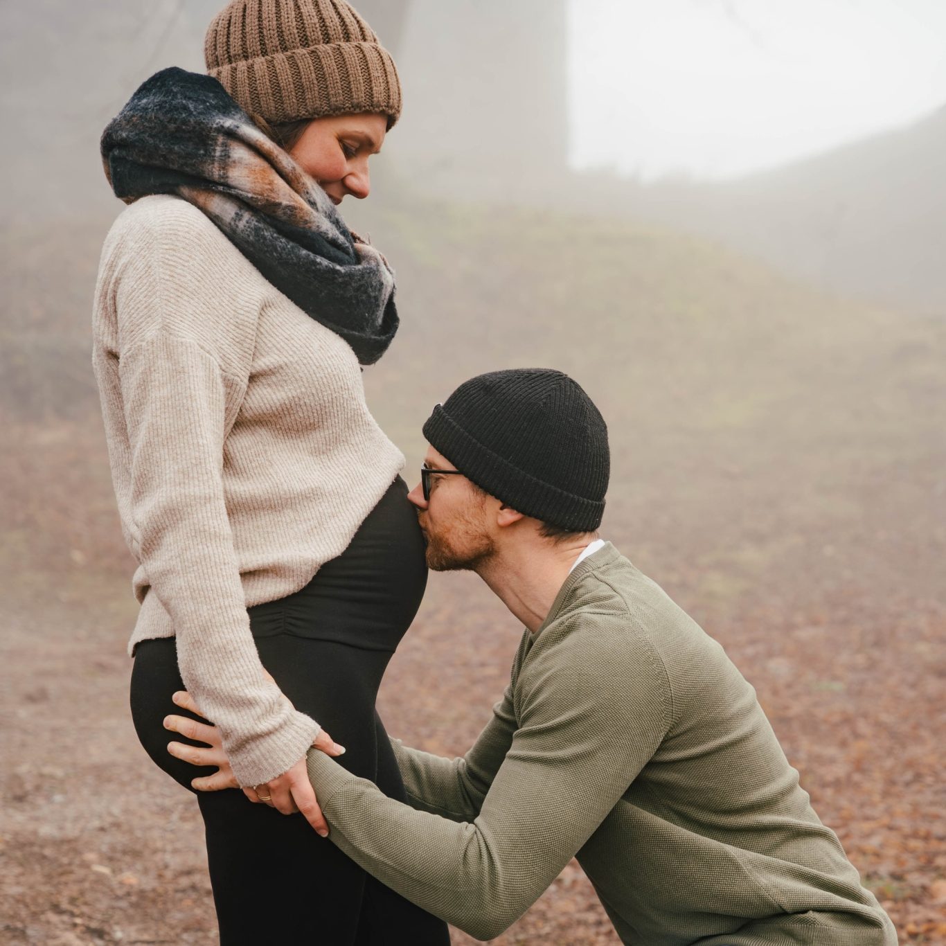 Schwangere Frau in warmer Kleidung, der Partner küsst ihren Bauch in nebliger Umgebung.