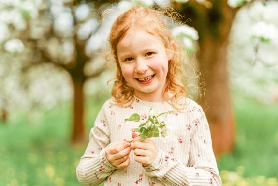 Lächelndes Mädchen mit lockigem Haar, hält Blumen in einem blühenden Obstgarten.