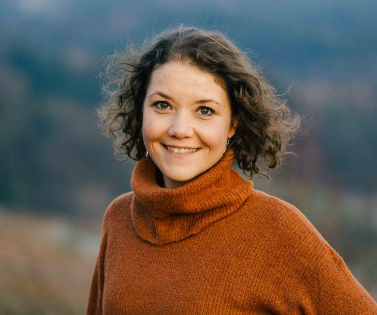 Fotografin mit lockigem Haar, lächelnd in orangefarbener Rolle steht vor verschwommenem Hintergrund.